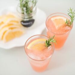 Refreshing pink lemonade drinks garnished with lemon slices and rosemary sprigs, served on a clean white surface.