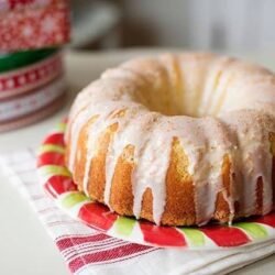 Golden vanilla bundt cake with glaze on festive holiday plate and wrapping paper in background.