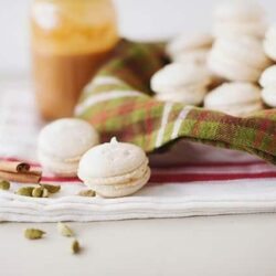 Delicate white macarons with a jar of caramel and cinnamon sticks on a white surface.