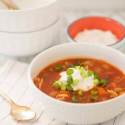 Creamy chicken enchilada soup with green onions in a white bowl on a white striped cloth.