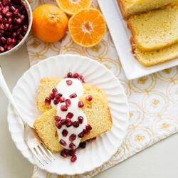 Creamy cake slices with pomegranate topping on a white plate, fresh fruit and cake slices in the background.