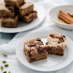 Fudgy chocolate chip cookie bars on white plate with cinnamon sticks and baked blondies in background.