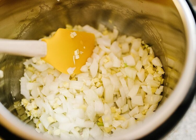 Chopped onions and garlic in a stainless steel mixing bowl with a yellow spatula.