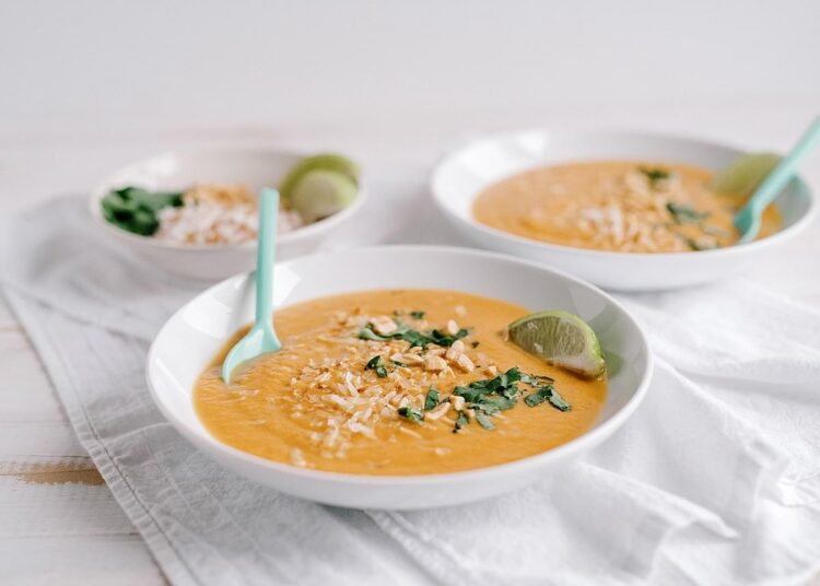 Creamy butternut squash soup garnished with chopped herbs and lime wedges, served in white bowls with pastel spoons.
