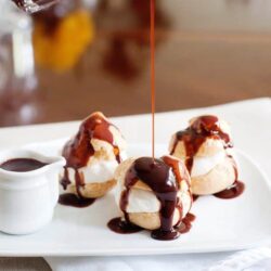 Vanilla ice cream profiteroles topped with rich chocolate sauce on white plate.