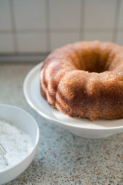 Butter Golden Bundt Cake on a white plate with vanilla glaze and powdered sugar in a white bowl.