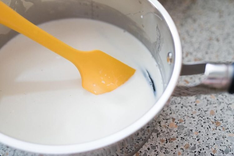 Creamy milk being heated in a stainless steel pot with a yellow spatula for baking or cooking.