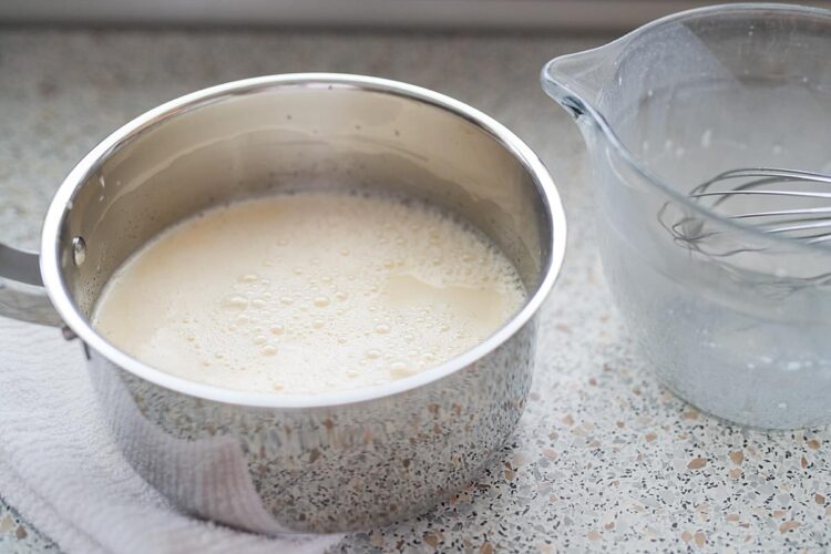 Creamy milk simmering in a stainless steel pot with a glass measuring pitcher and whisk on terrazzo countertop.