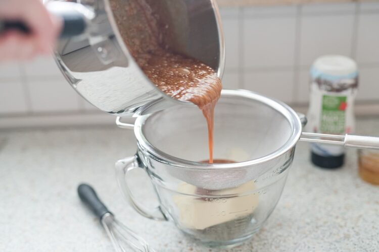 Rich chocolate mixture being poured through a fine sieve into a measuring cup for baking.