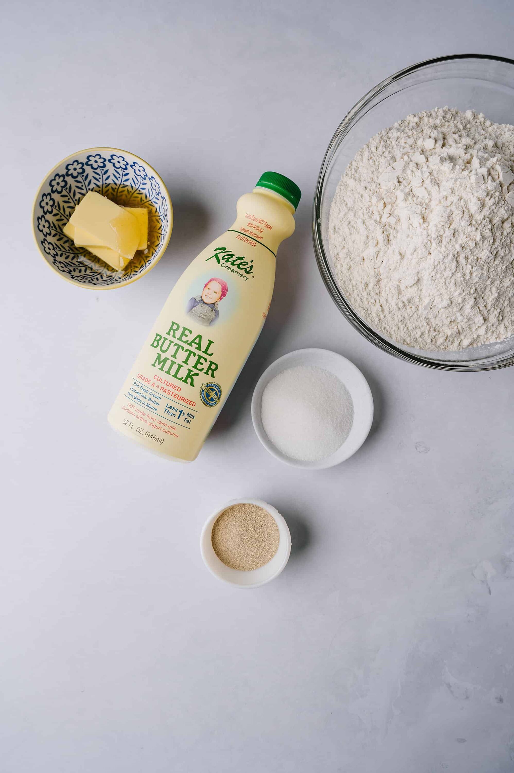 Flour, butter, sugar, and milk ingredients for baking on light gray background.