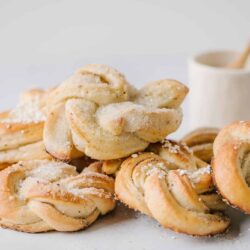 Sweet cinnamon knots pastry with powdered sugar and a small jar of whipped cream on white background.