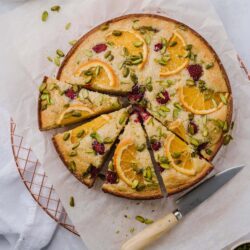 Fresh fruit upside-down cake topped with orange slices, raspberries, and pistachios on a wire rack.