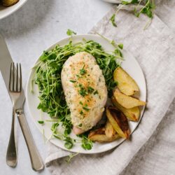 Creamy baked chicken breast topped with herbs served with roasted potatoes and fresh microgreens on a white plate.