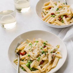 Creamy chicken pasta with peas and sun-dried tomatoes, served in white bowls with glasses of water.