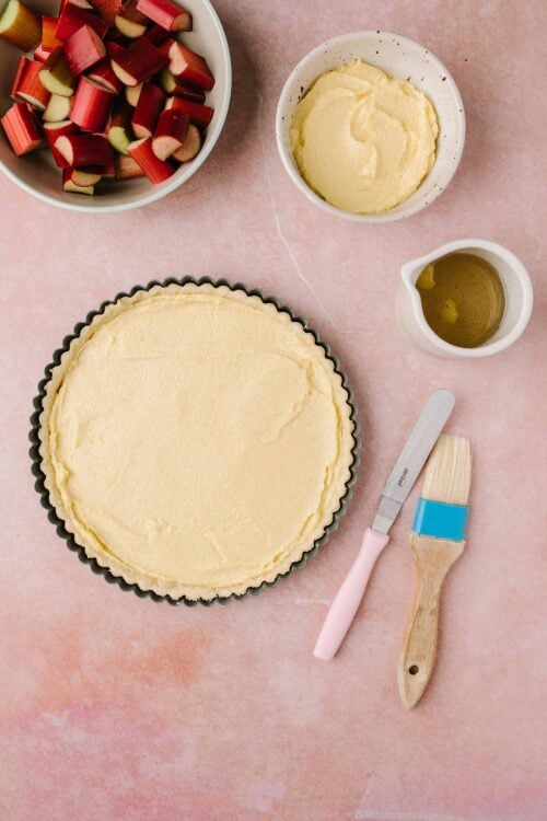 Buttery pie crust dough in a tart pan with rhubarb and custard ingredients around it, ready for baking.