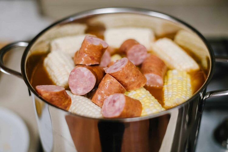 Sauteing sliced sausage and corn on the cob in a stainless steel skillet with broth.