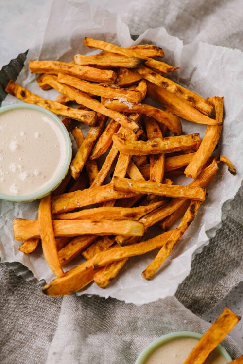 Golden baked sweet potato fries with a side of creamy dipping sauce, served on parchment paper. Perfect for healthy snacking or dinner.