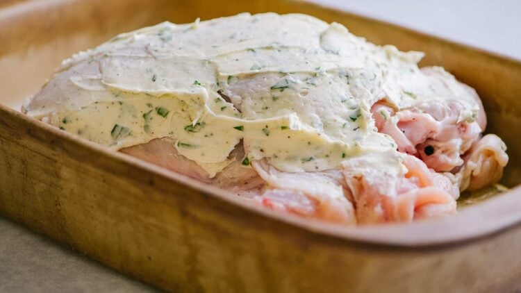 Creamy herb sauce spreads over raw chicken in a wooden baking dish.
