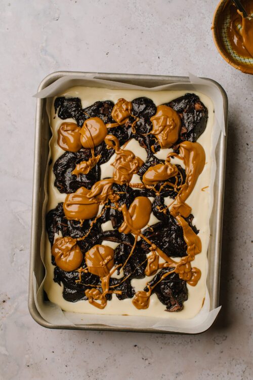 Rich chocolate brownie layered with white cream and topped with caramel sauce, in a baking dish.