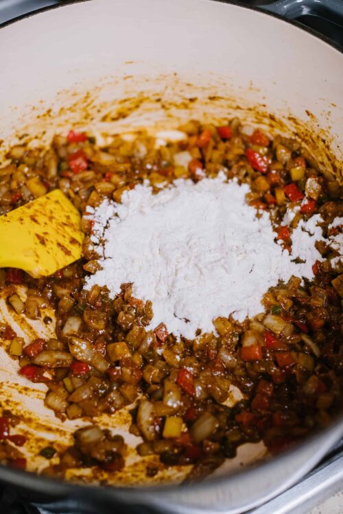 Savory vegetable and meat mixture cooking in a pot, with flour added for baking preparation.