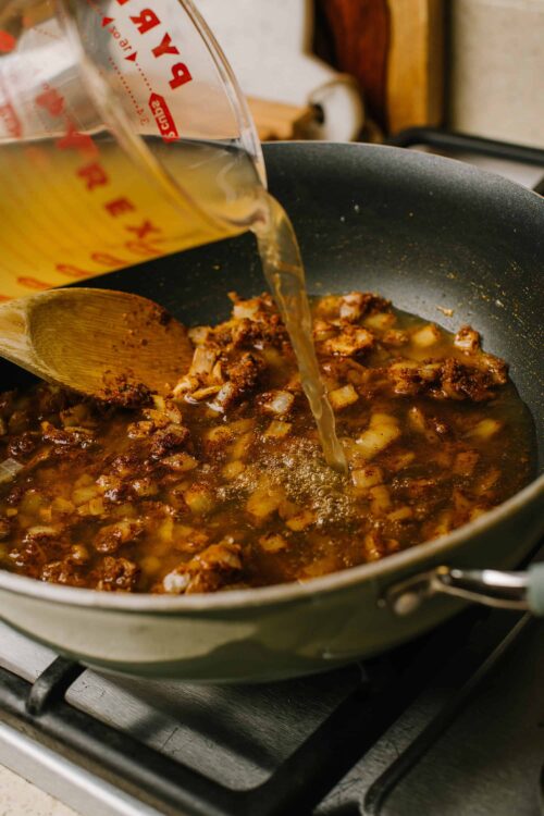 Beef stew simmering in a skillet with broth pouring in from measuring cup.