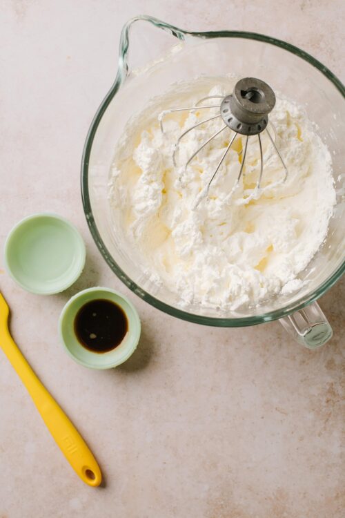 Fluffy whipped cream in a glass mixing bowl with a hand mixer attachment, on a light countertop.