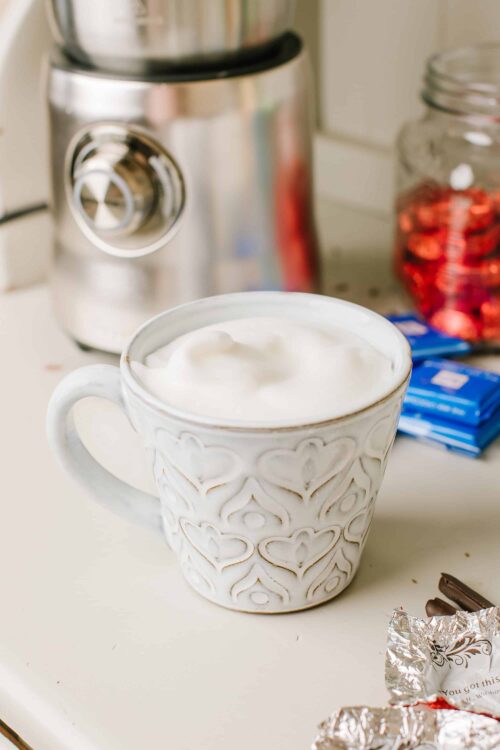 Frosted mug of whipped cream or hot chocolate on a kitchen countertop with chocolates and a blender in the background.