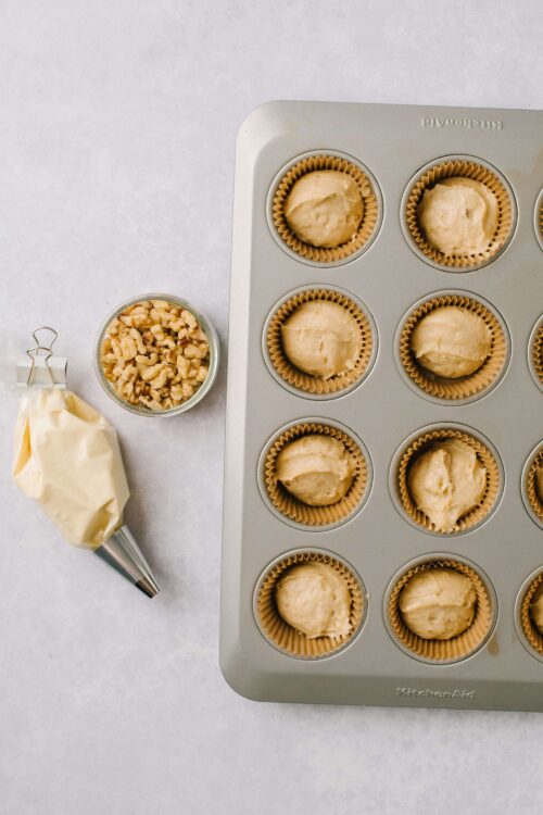 Butterscotch blondie batter in cupcake liners in a baking tray with a piping bag and chopped nuts on the side.