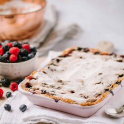 Homemade mixed berry crumble pie with powdered sugar on top, fresh berries, and a rustic kitchen setting.