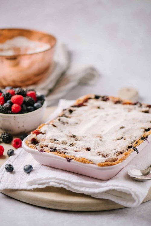 Homemade mixed berry crumble pie with powdered sugar on top, fresh berries, and a rustic kitchen setting.