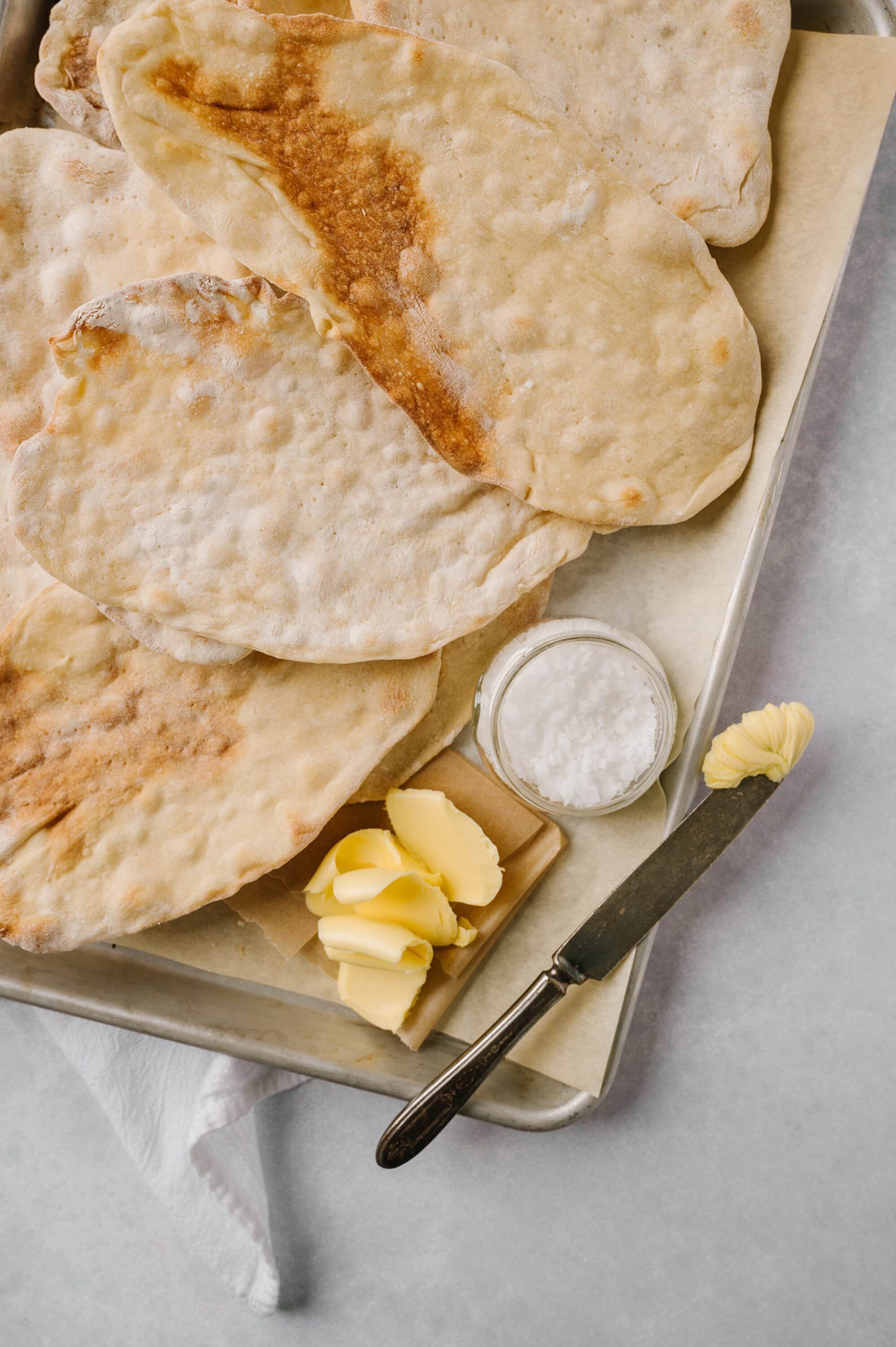 Fluffy homemade naan bread with butter, salt, and a butter knife on a baking sheet.