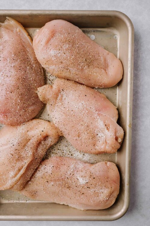 Raw chicken breasts seasoned with salt, pepper, and spices on a baking sheet for oven roasting.