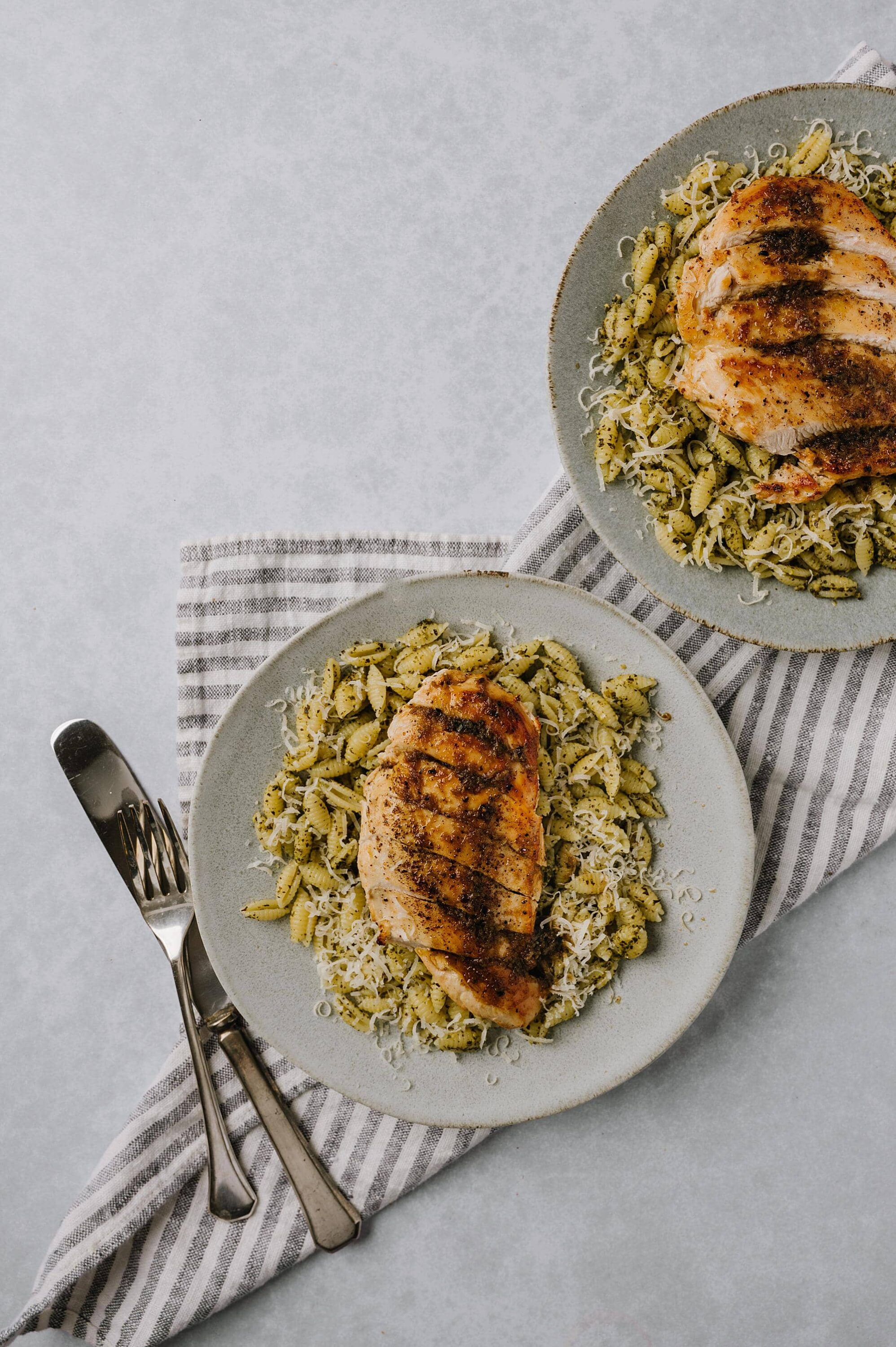 Seared chicken breast with pesto pasta and grated cheese on gray plates.