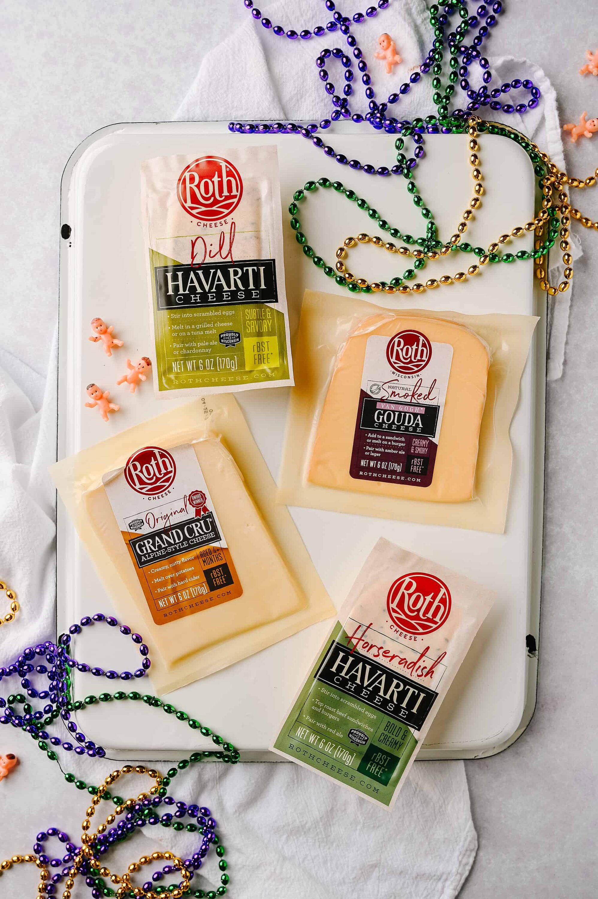Roth cheese varieties on a white tray with colorful beads and mini baby figurines, celebrating Mardi Gras or festive occasion.