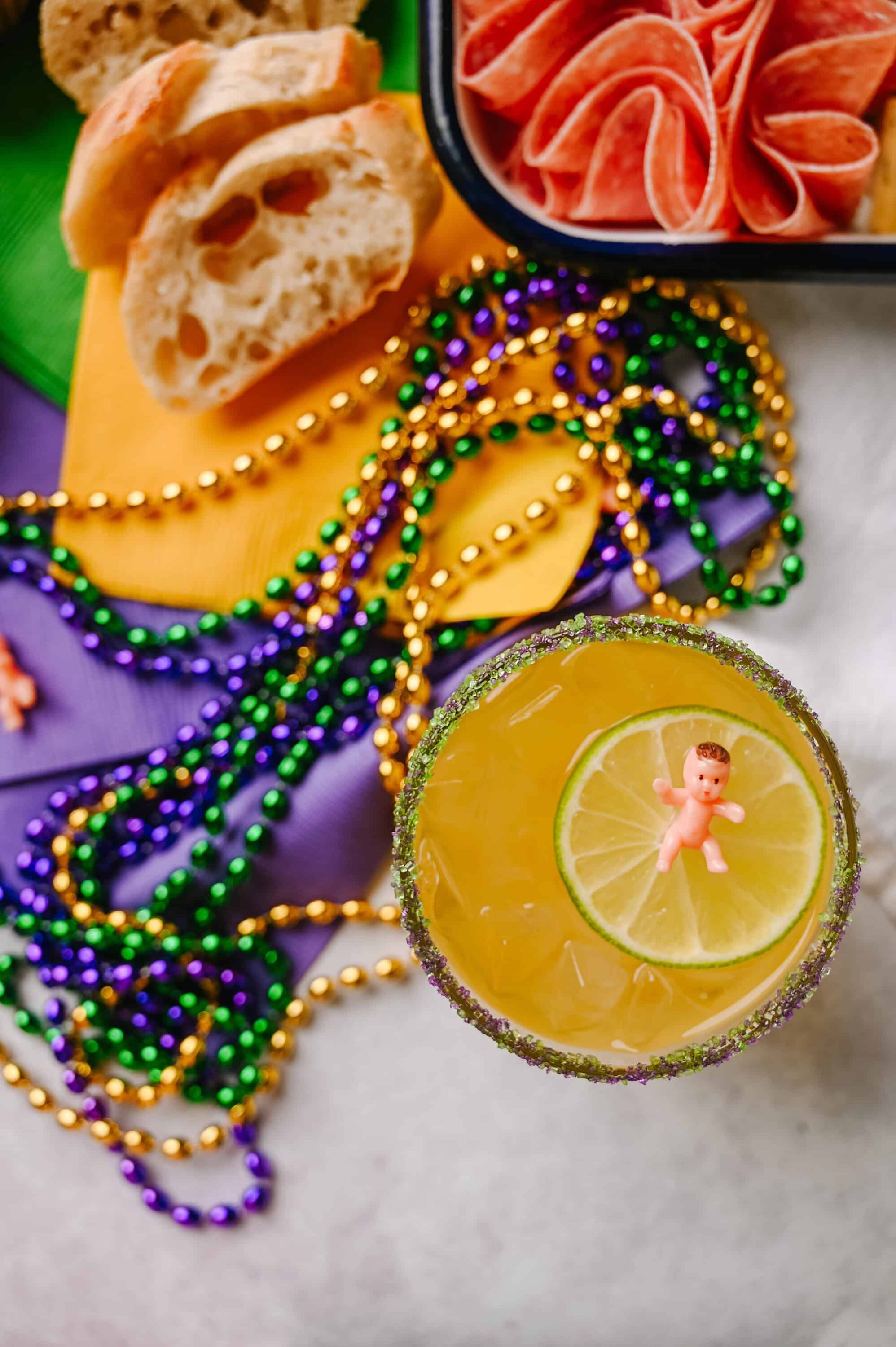 Beaded Mardi Gras beads, lemon cocktail with a baby figurine, and festive snacks for celebration.