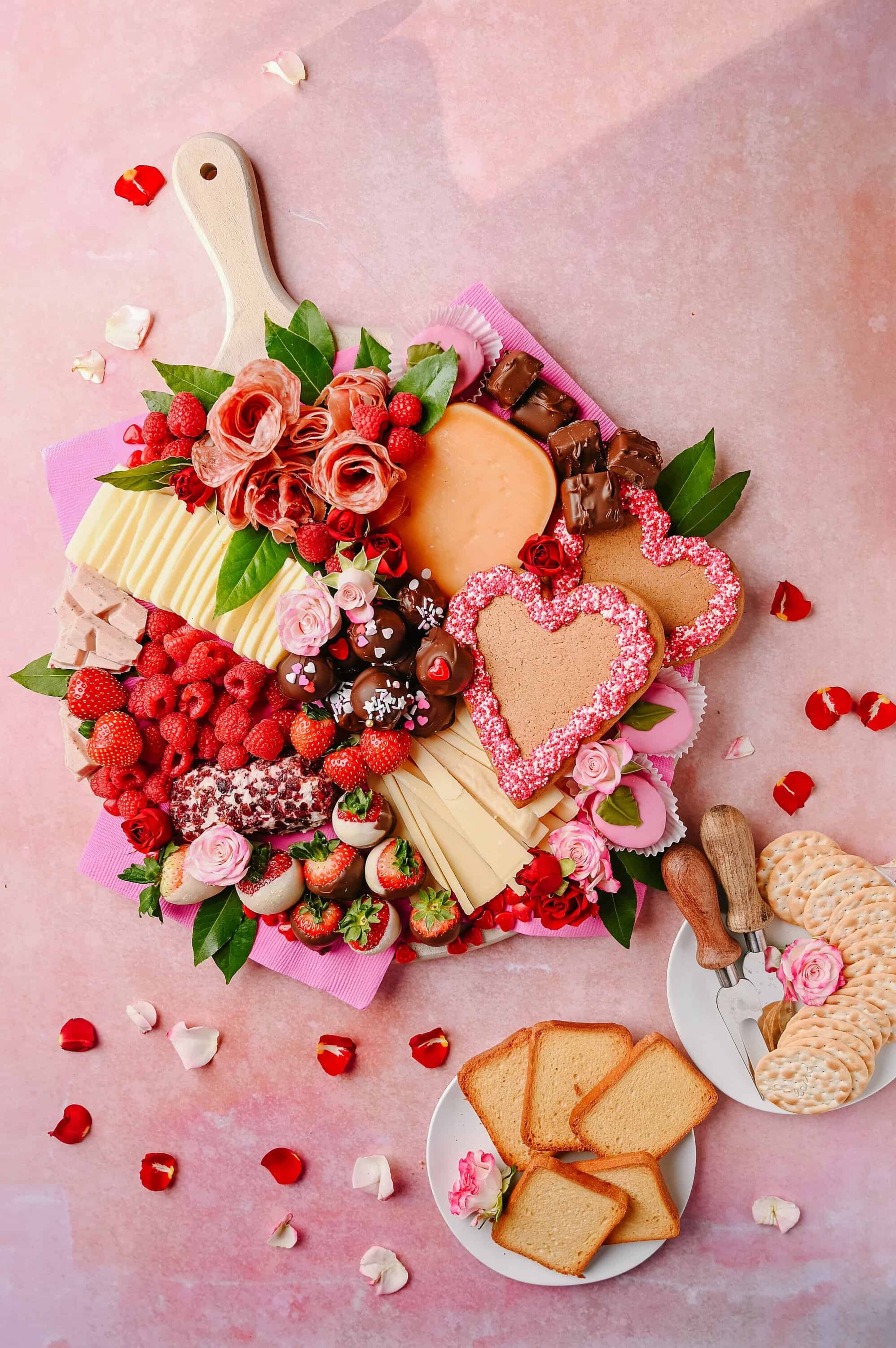 Colorful charcuterie and dessert board with strawberries, chocolates, cheese, and heart-shaped cookies on pink background.