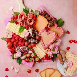 Colorful charcuterie and dessert board with strawberries, chocolates, cheese, and heart-shaped cookies on pink background.