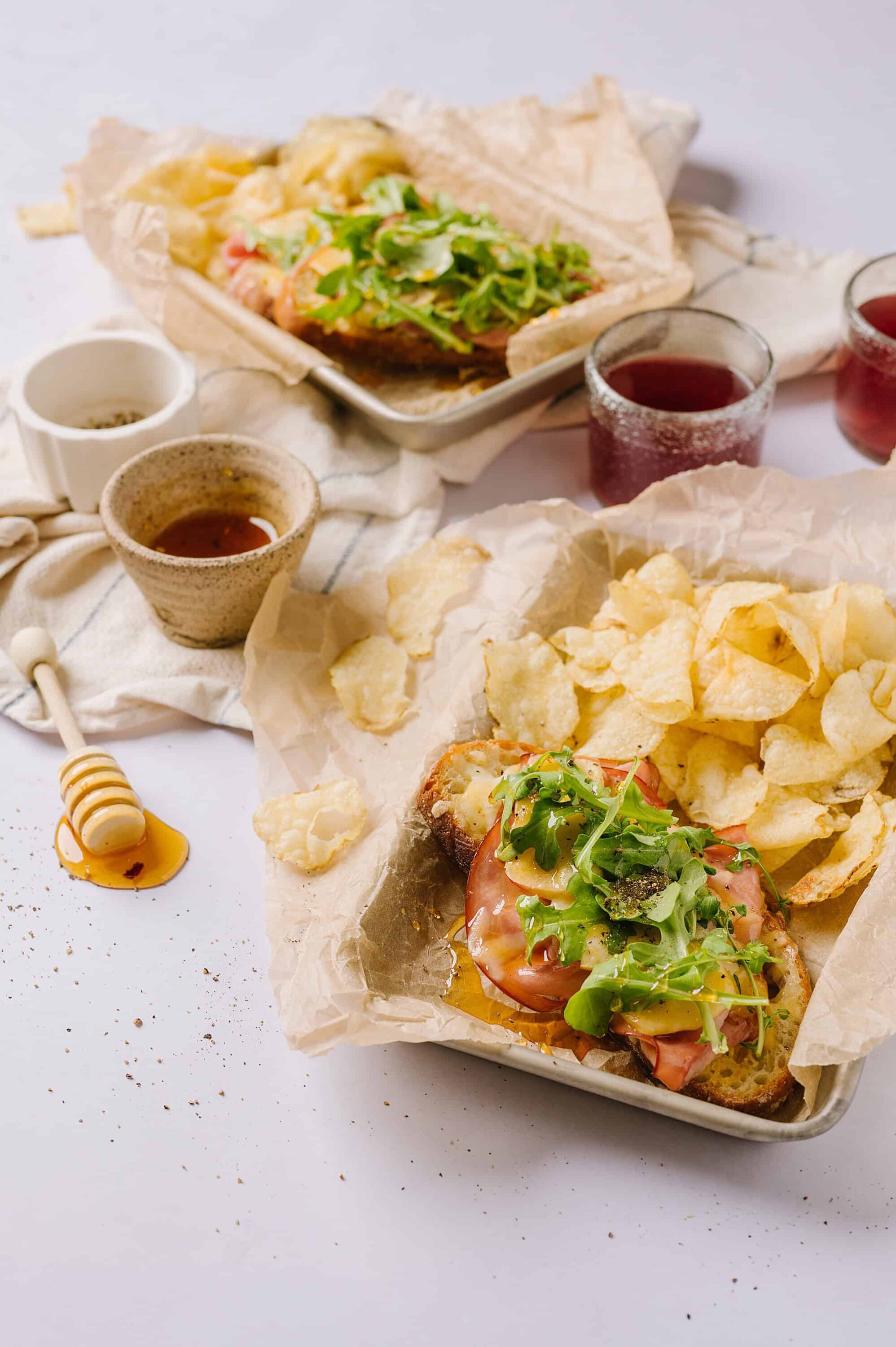 Fresh homemade open-faced turkey sandwich with greens and chips on parchment paper, with honey and drinks on a white background.