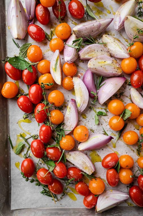 Vibrant cherry tomatoes, red onion, and fresh herbs on baking sheet with olive oil drizzle.