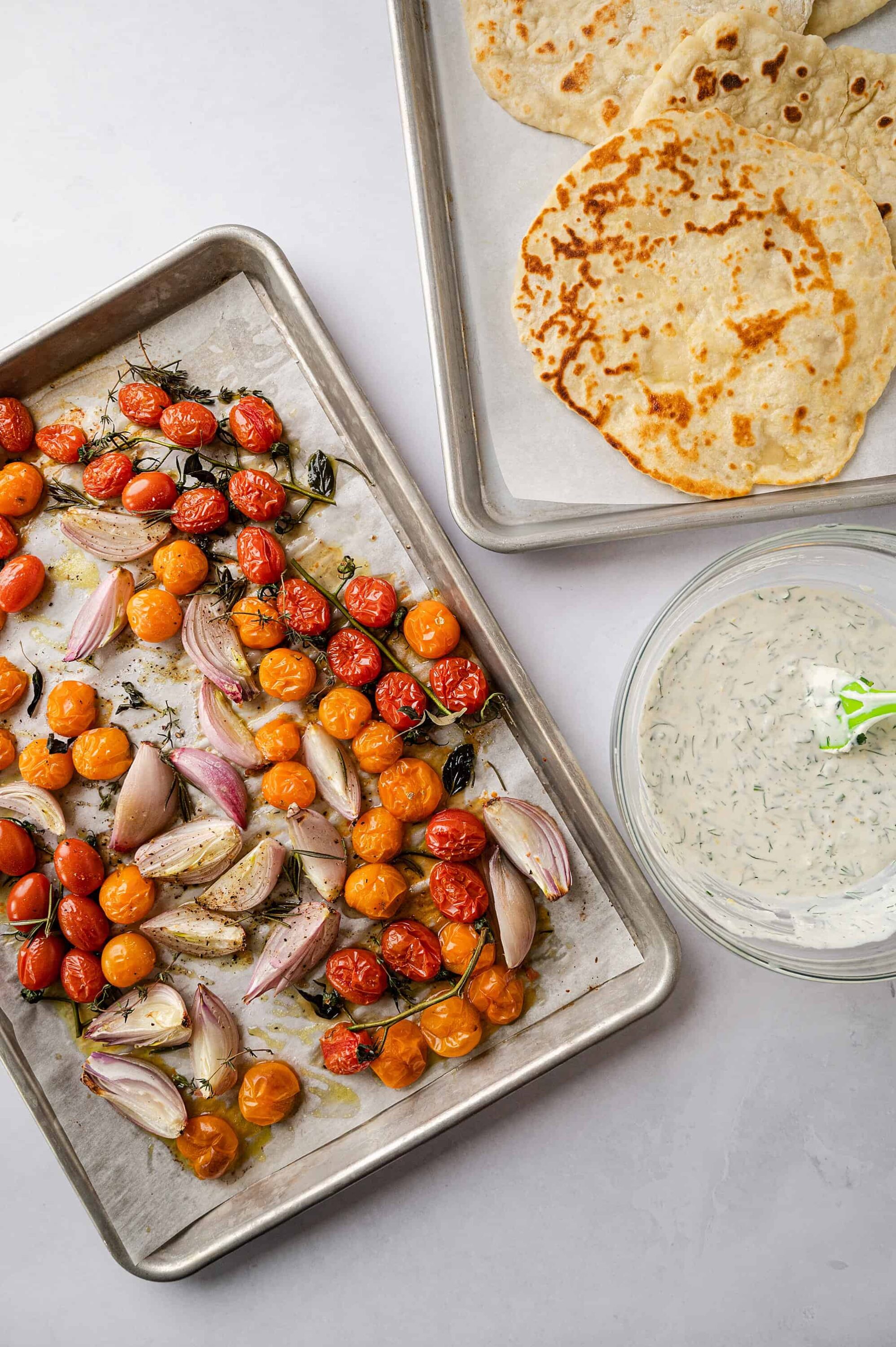 Roasted cherry tomatoes and garlic on a baking sheet with flatbreads and herbed tzatziki sauce on the side.