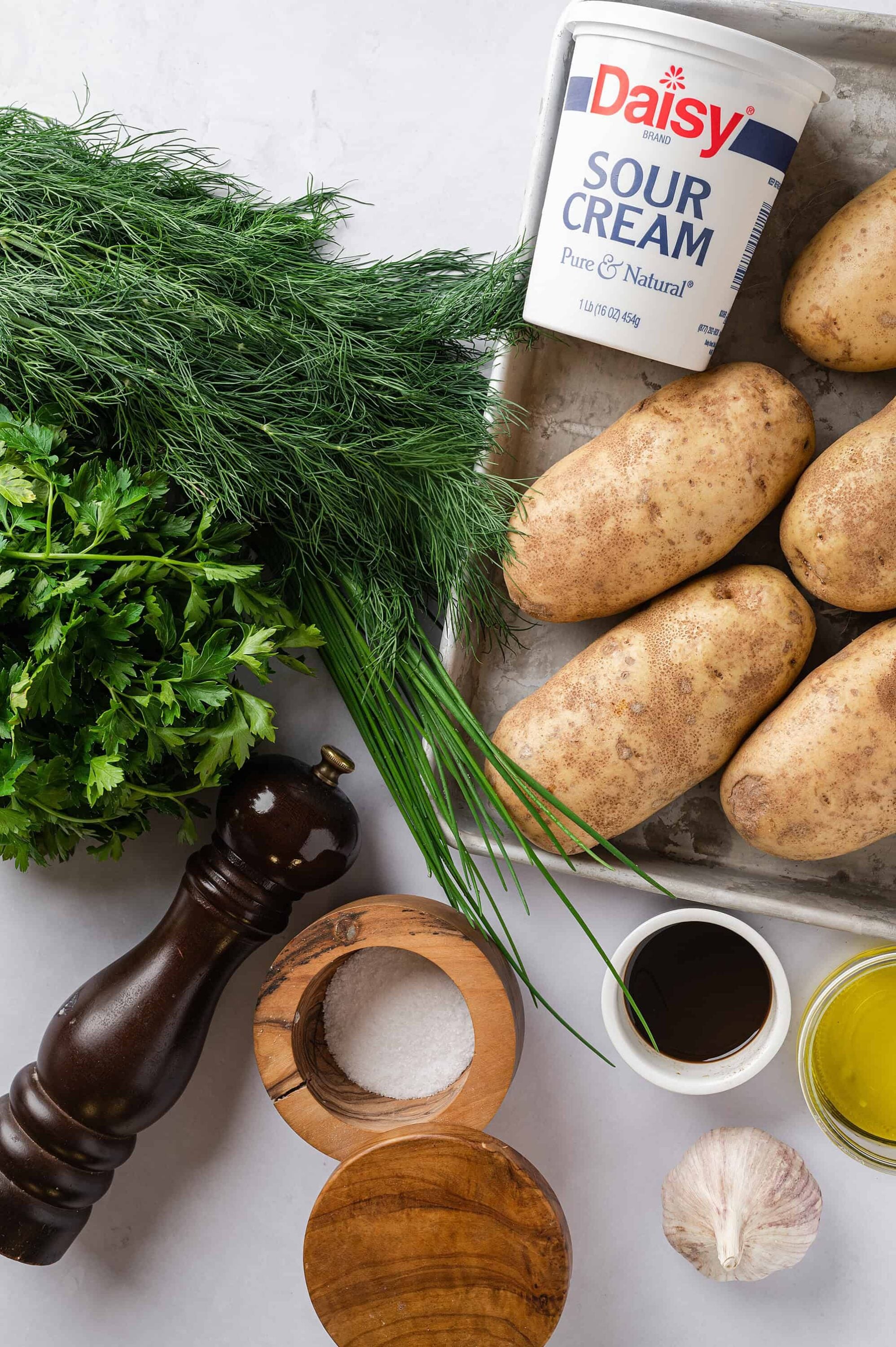Fresh herbs, potatoes, sour cream, garlic, olive oil, salt, pepper grinder for homemade baked potato recipe.