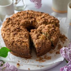 Cinnamon Apple Coffee Cake with crumb topping on a white plate with lilacs and coffee cups in the background.