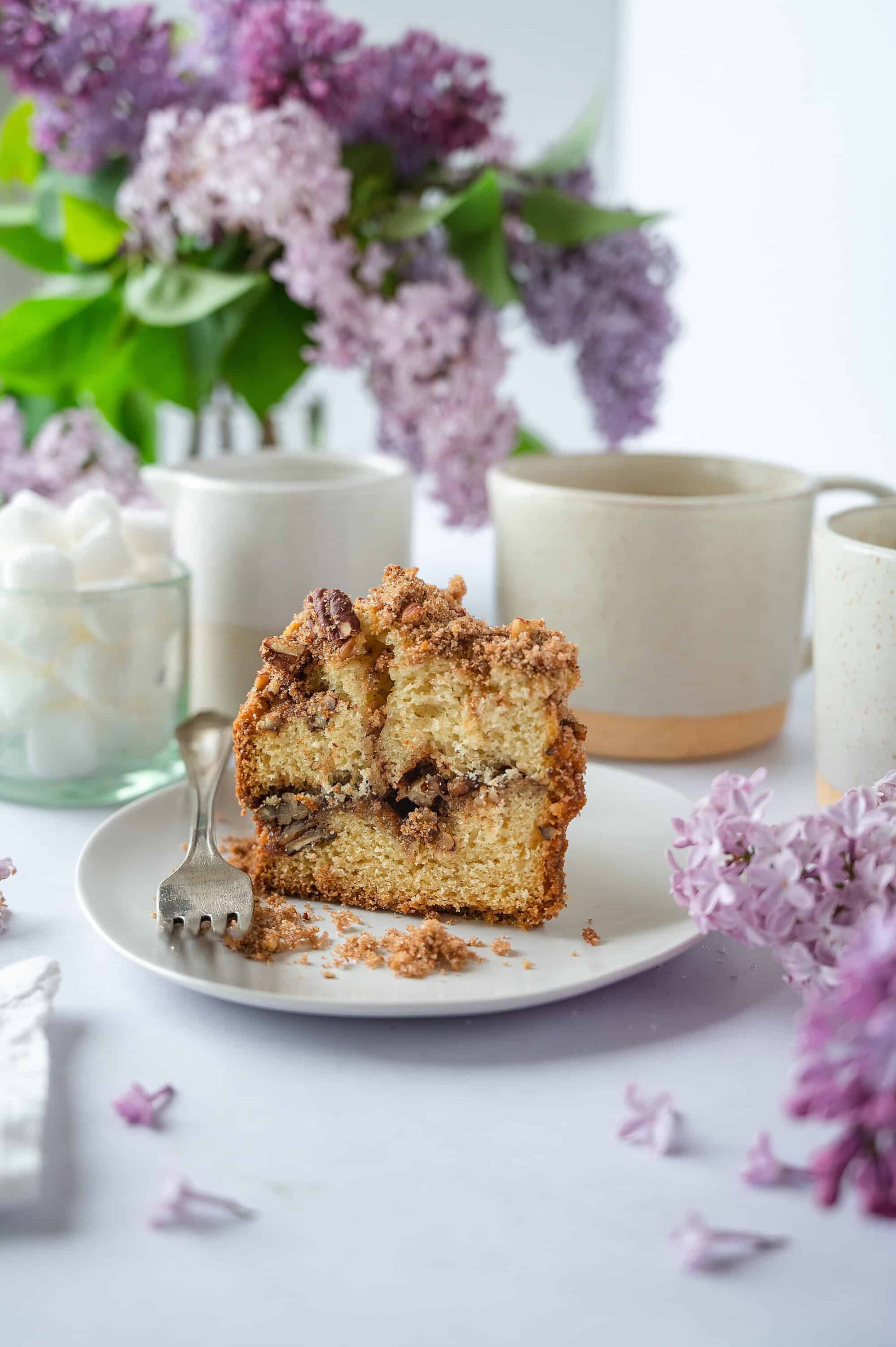 Butter cake with chocolate chips on a white plate surrounded by lilac flowers and tea cups, bakedbree.com.