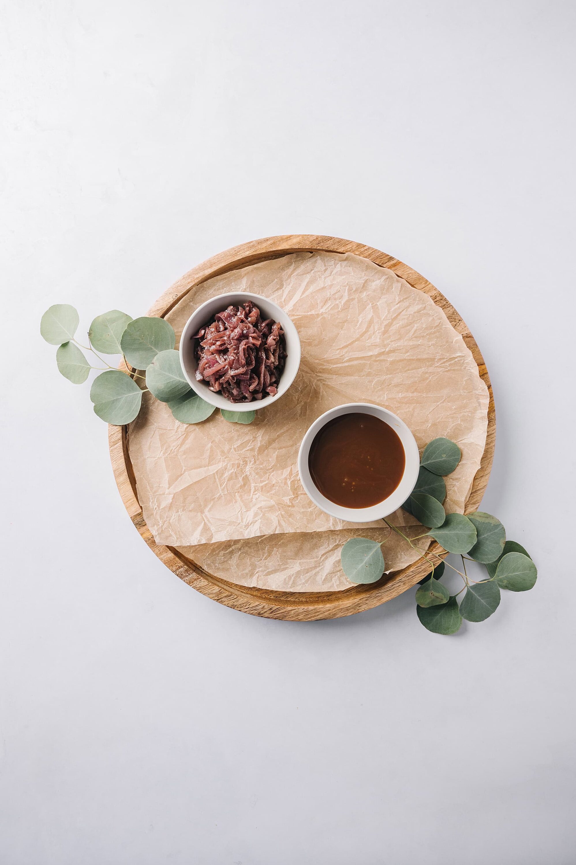 Sliced beef and dipping sauce on wooden platter with eucalyptus leaves, minimal food presentation, clean white background, modern culinary style.