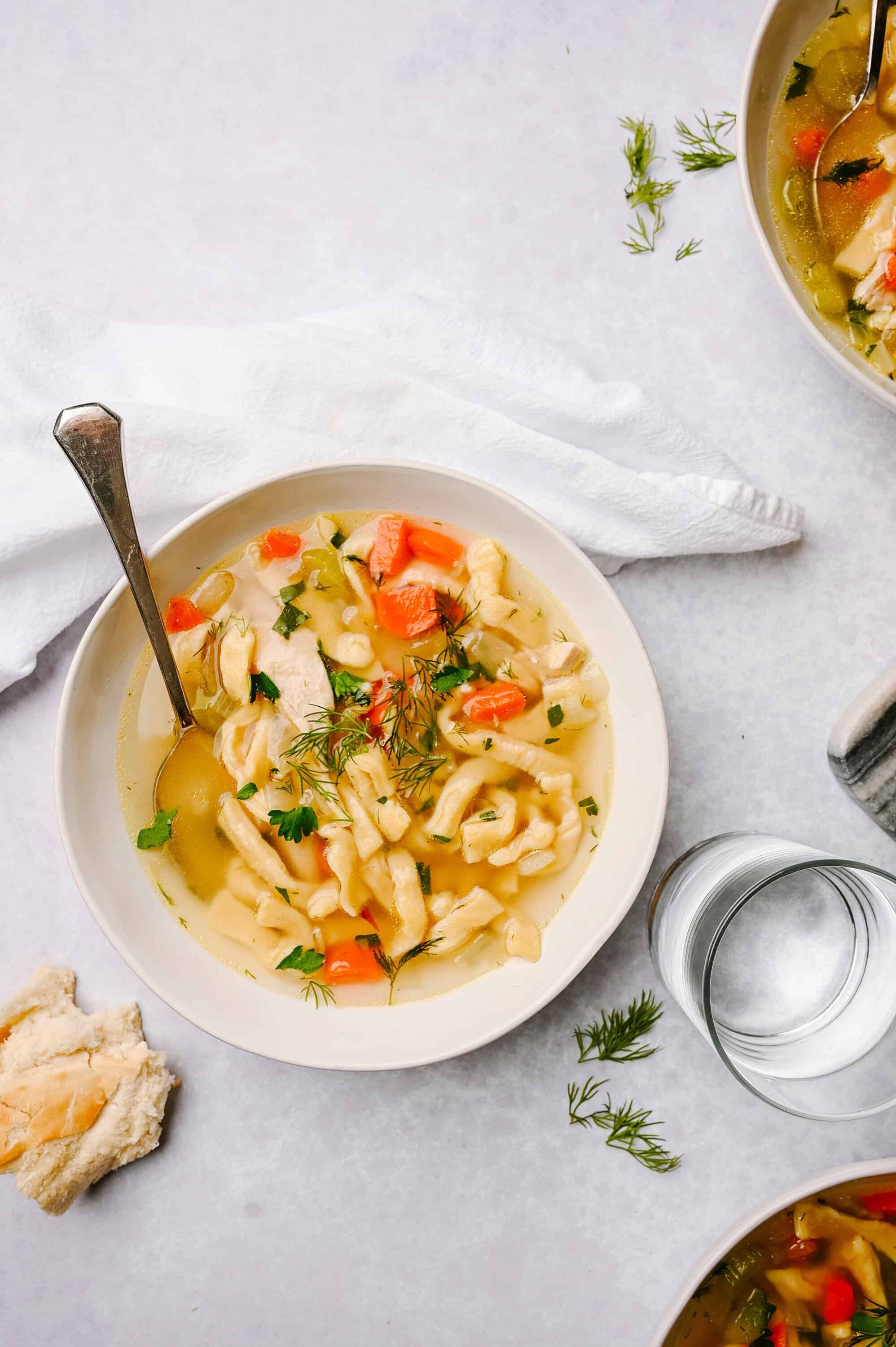 Creamy chicken noodle soup with vegetables, fresh herbs, and a slice of bread on a white background.