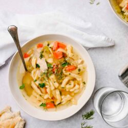 Creamy chicken noodle soup with vegetables, fresh herbs, and a slice of bread on a white background.