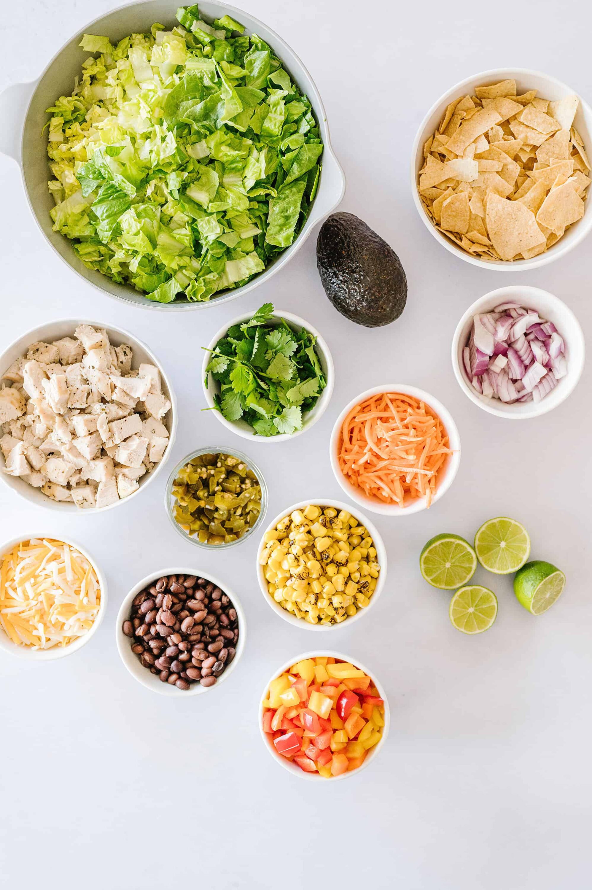 Fresh vegetable ingredients for taco salad, including lettuce, corn, chicken, avocado, diced onions, and shredded cheese.