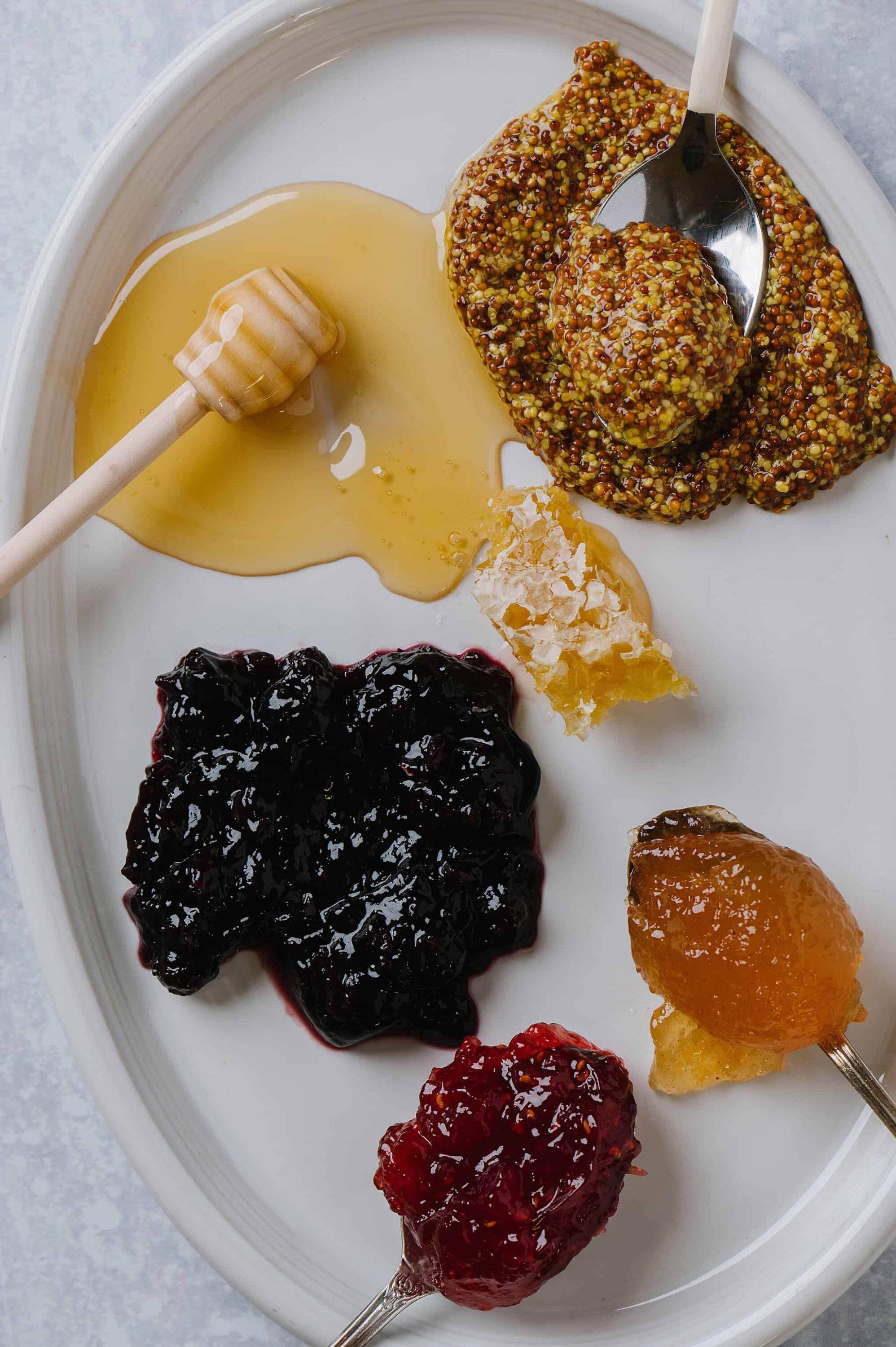 Honey on a stick with honeycomb and an assortment of fruit preserves on a white plate.