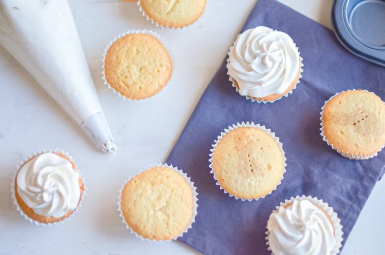 Freshly baked vanilla cupcakes with swirled white frosting on a white and blue background.