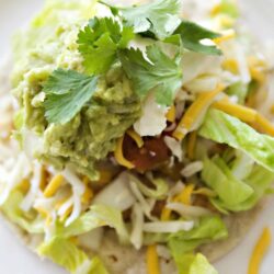 Fresh taco with guacamole, shredded cheese, lettuce, and cilantro on a white plate.
